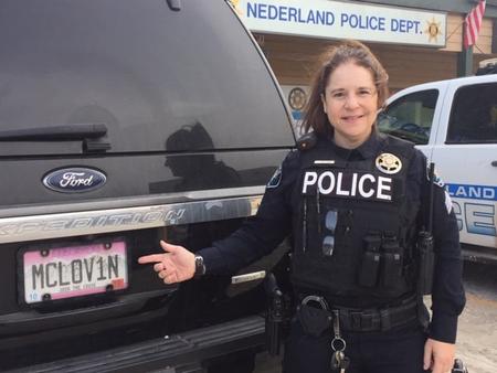 Sergeant Jennifer Fine-Loven standing next to a Nederland Patrol Car, pointing at the license plate, which reads MCL0V1N